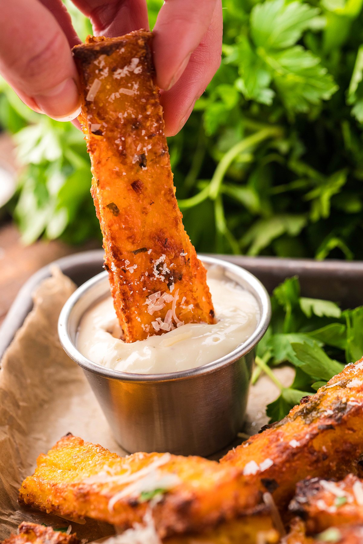A hand dips a crispy cornmeal french fry topped with cheese into a cup of creamy truffle mayo dipping sauce, with herbs in the background.