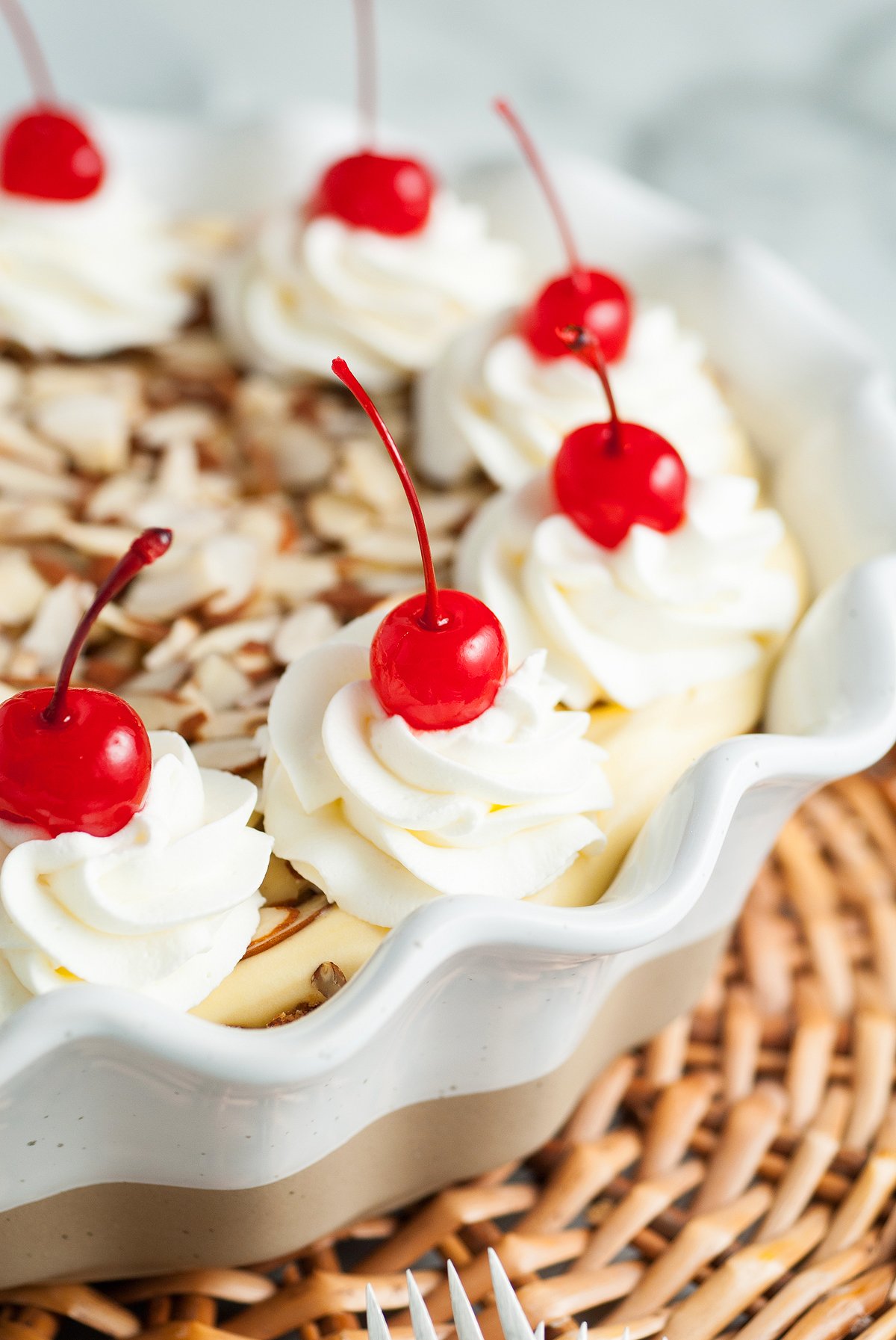 A pie with an almond crust, topped with whipped cream swirls, maraschino cherries, and sliced almonds in a decorative dish—a truly Italian inspired dessert.