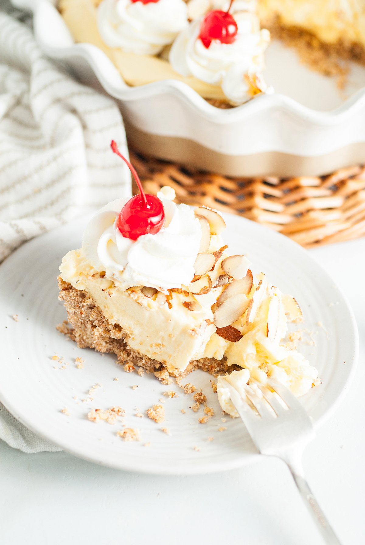 A slice of creamy Almond amaretto pie topped with whipped cream, almond slices, and a cherry sits on a white plate with a fork—an Italian inspired dessert to savor.