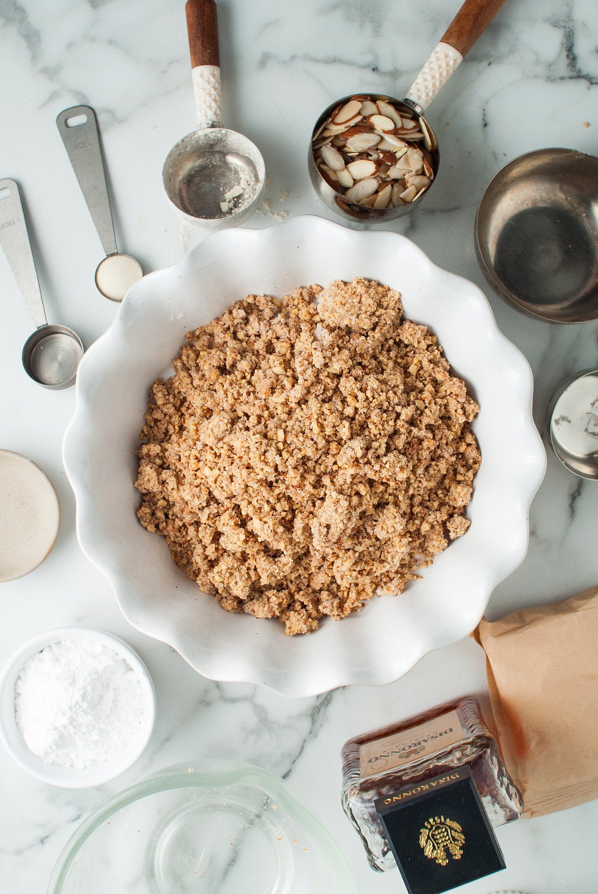 A pie dish with crumbly topping, surrounded by baking tools, sliced almonds, and ingredients on a marble counter—perfect for creating an Italian inspired dessert like almond amaretto pie.