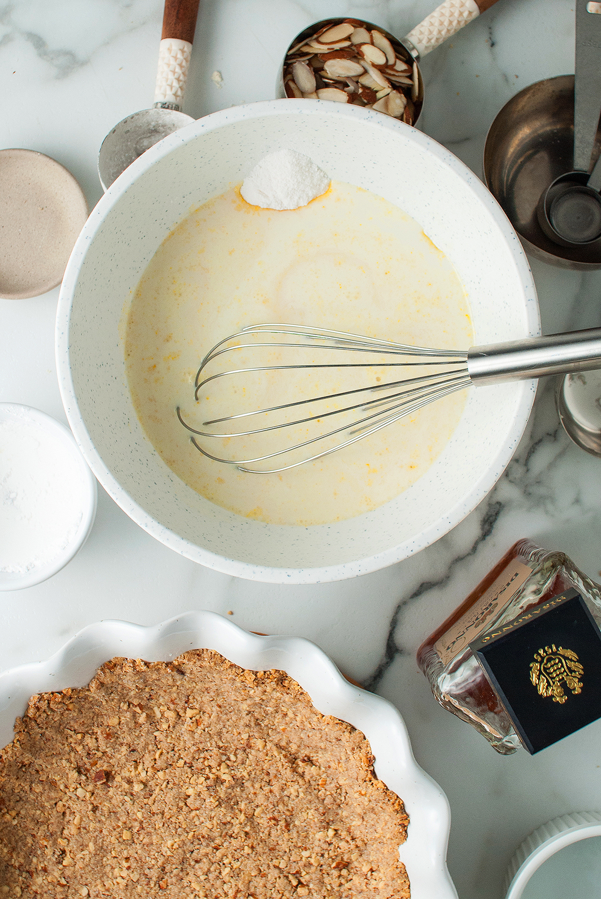 A bowl with batter and a whisk sits next to an almond crust, cinnamon, and baking ingredients on a countertop—perfect for preparing a delicious almond amaretto pie.