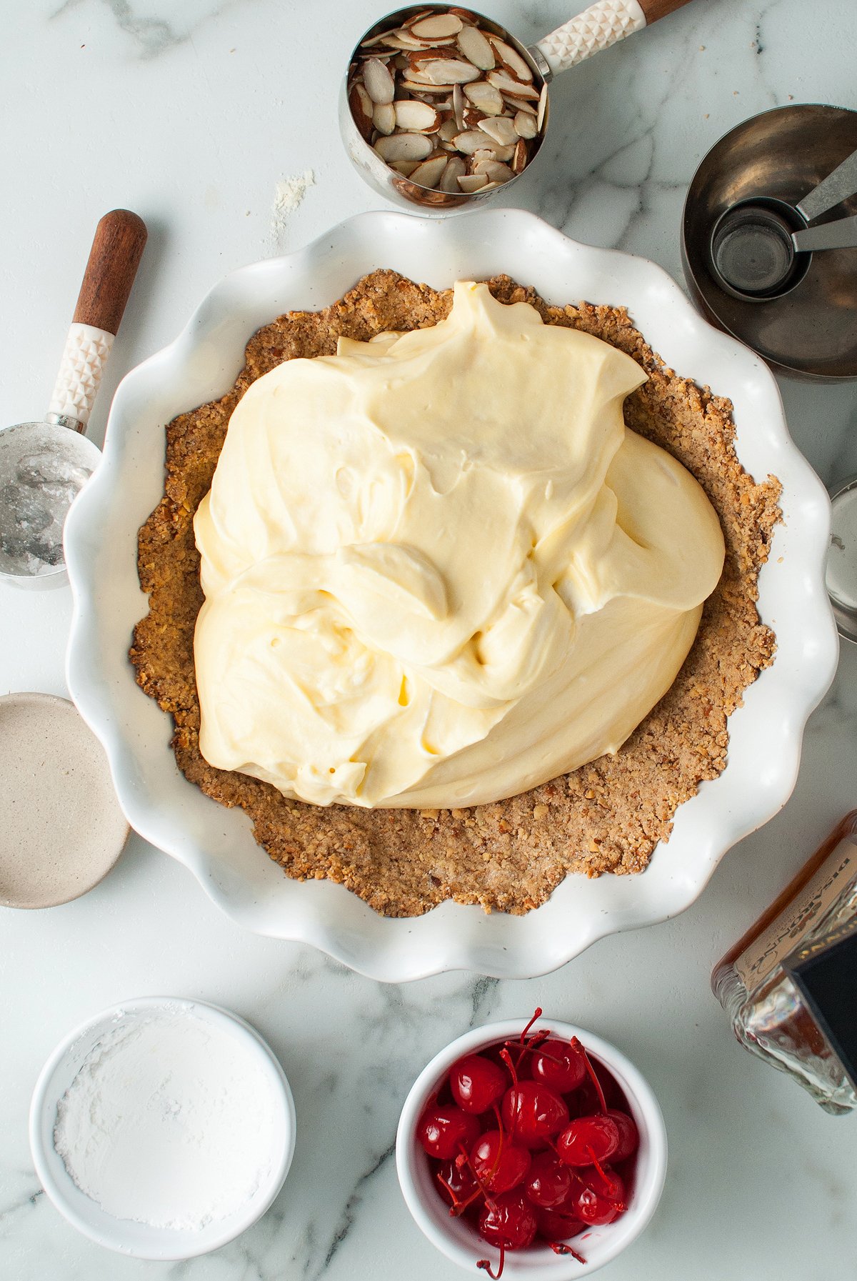 Overhead view of an almond crust filled with creamy yellow filling, surrounded by baking tools and ingredients—an Italian inspired dessert ready for baking.