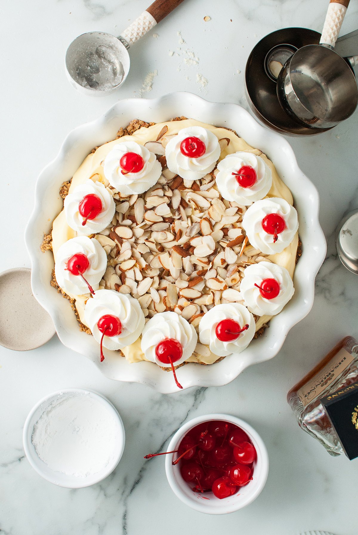 An Almond amaretto pie topped with whipped cream, cherries, and sliced almonds sits on a marble counter with baking tools.