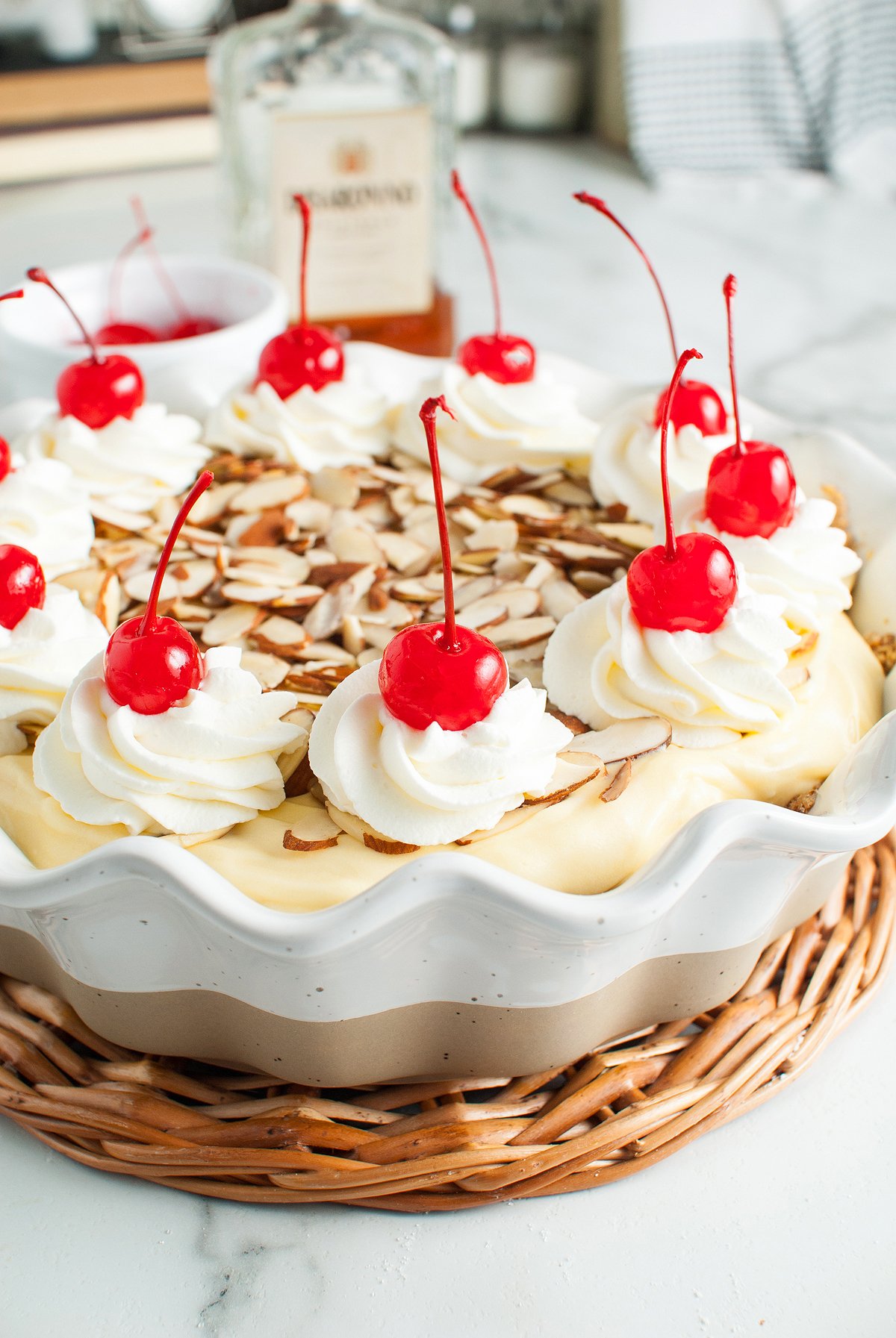 Creamy Amaretto Cream Pie topped with whipped cream, cherries, and sliced almonds in a white dish on a wicker tray, featuring a rich almond crust for the perfect dessert with amaretto liqueur.