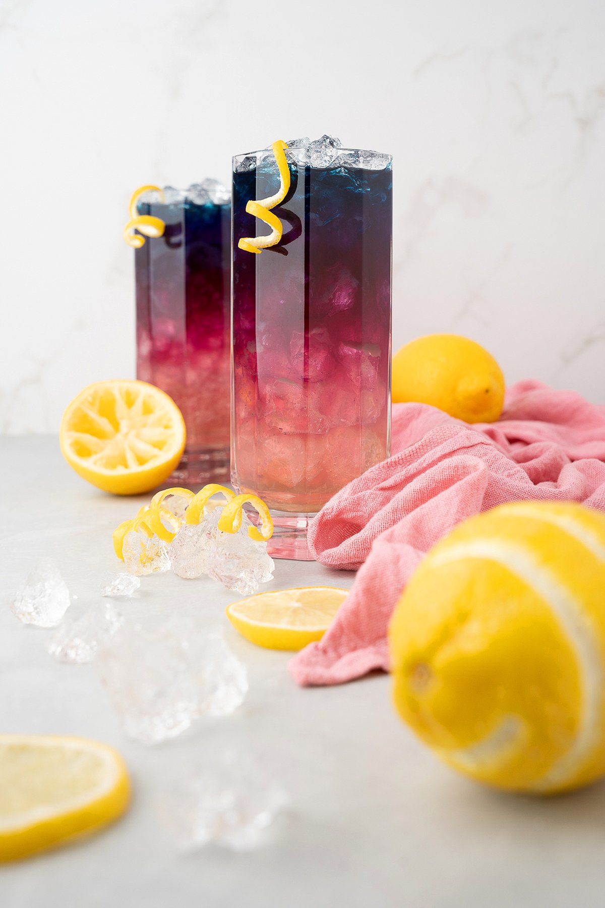 Two colorful layered cocktails with butterfly pea, ice, lemon twists, and sliced lemons rest on a light surface with a pink cloth—perfect for fans of color changing lemonade.