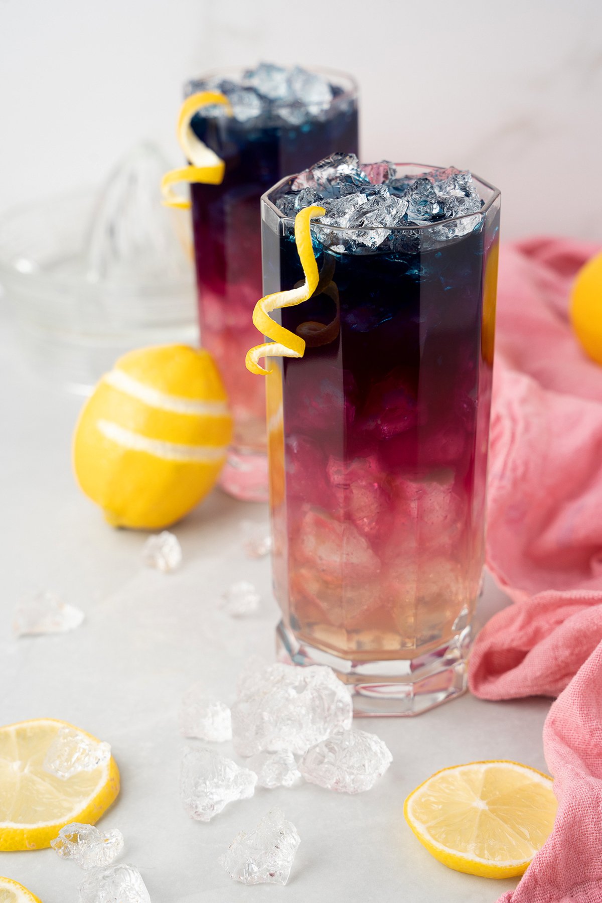 Two colorful layered cocktails featuring butterfly pea and ice with lemon twists, surrounded by fresh lemons and ice cubes on a white surface.