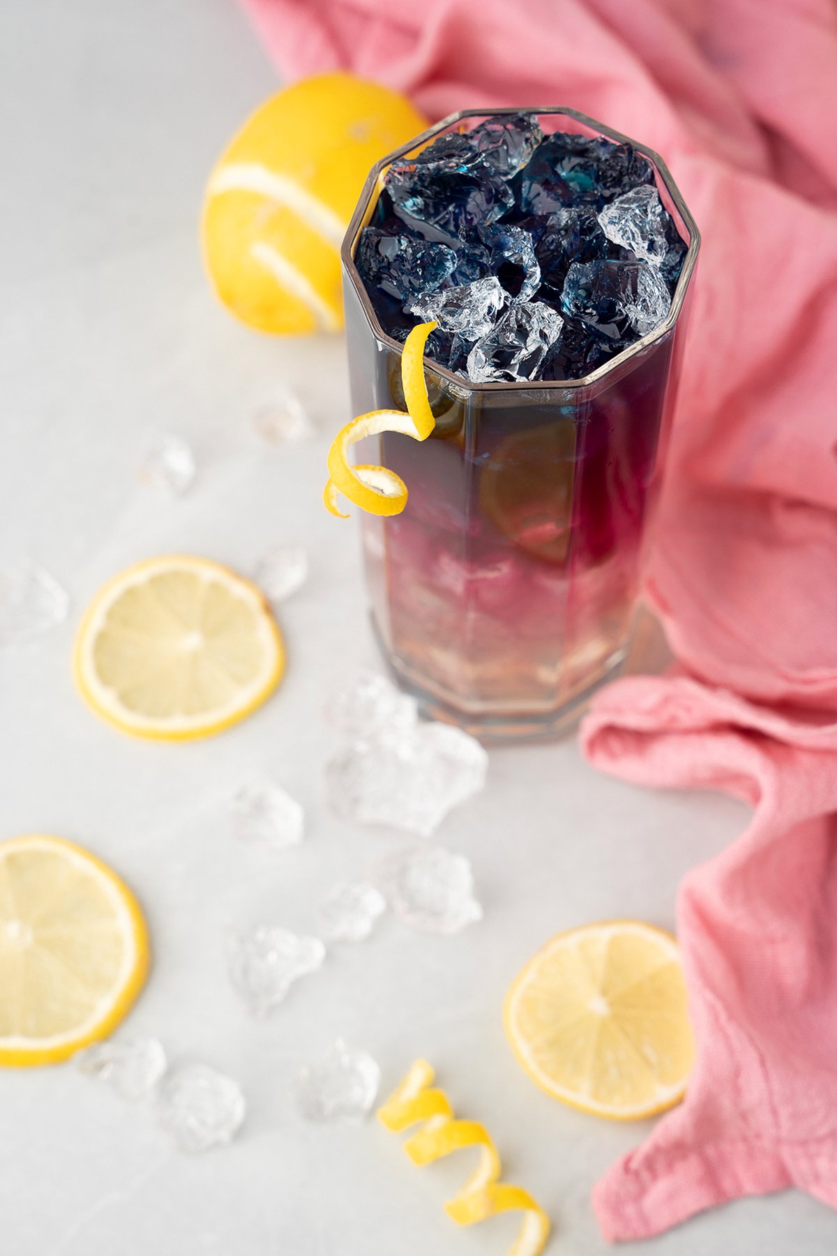 A tall glass of iced color changing lemonade made with butterfly pea, garnished with lemon and surrounded by lemon slices, ice, and a pink cloth.