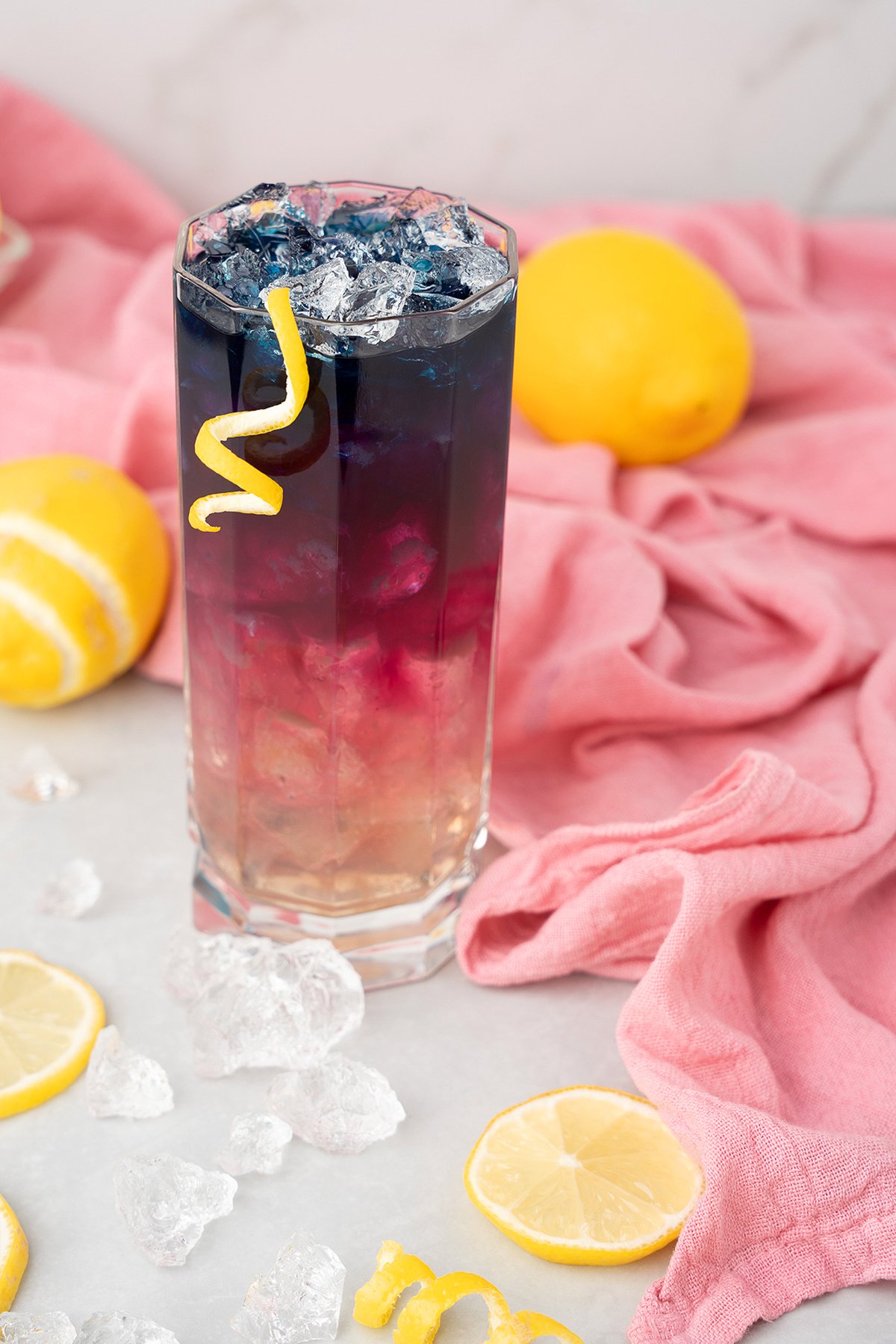 A layered cocktail with ice and butterfly pea, garnished with a lemon twist, surrounded by lemons and pink fabric.
