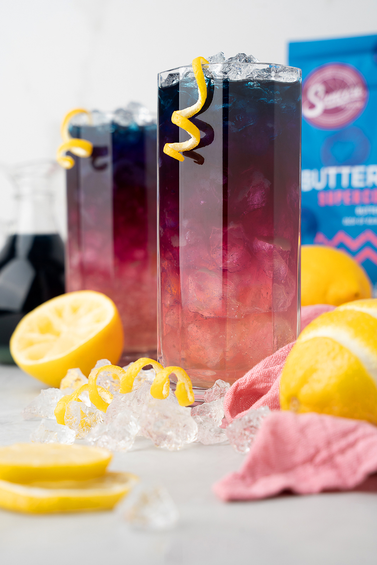 Two tall glasses with layered blue and pink butterfly pea color changing lemonade, garnished with lemon twists, sit among fresh lemons and ice.