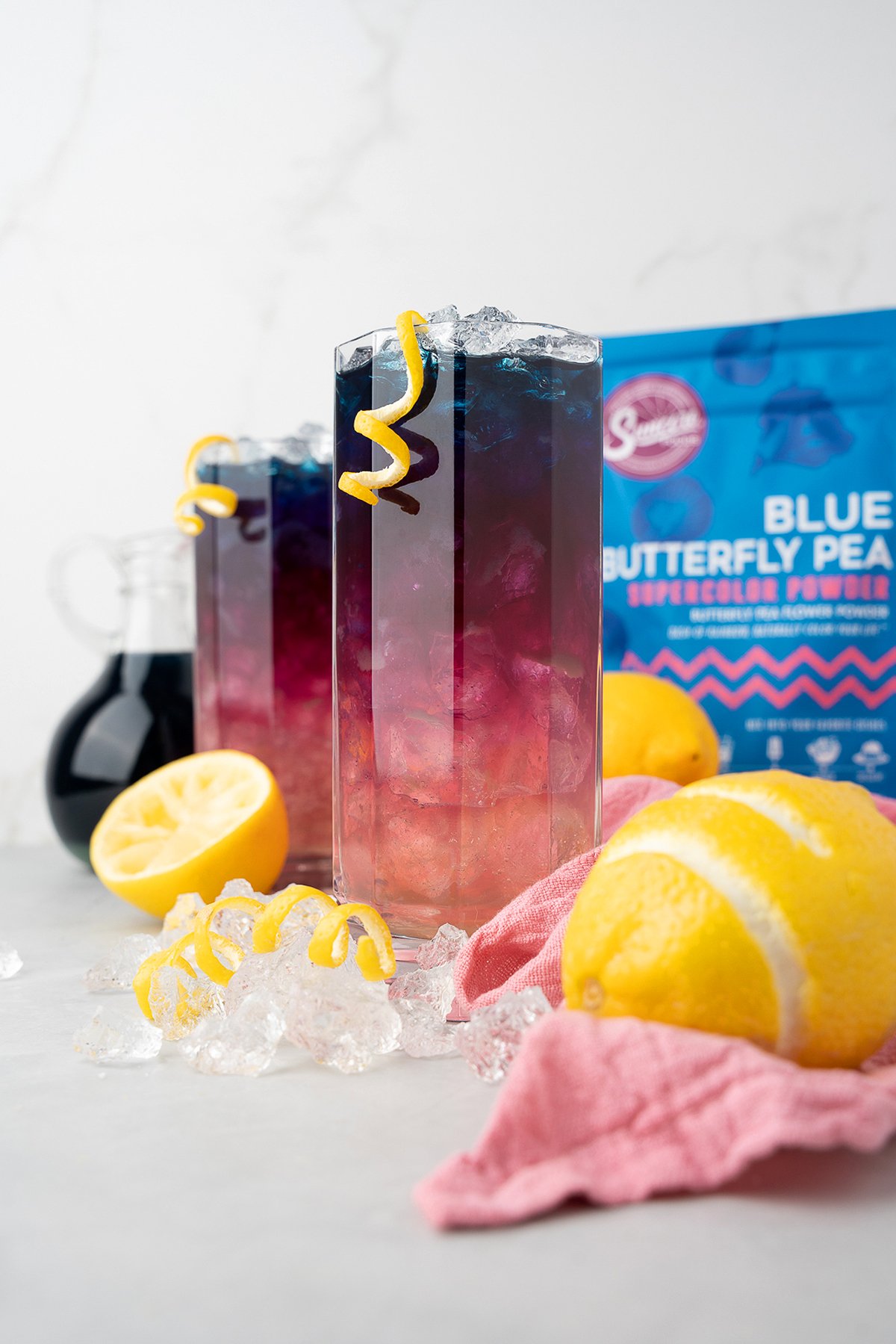 Tall glasses of colorful layered color changing lemonade with ice, lemon twists, and butterfly pea powder, surrounded by fresh lemons.