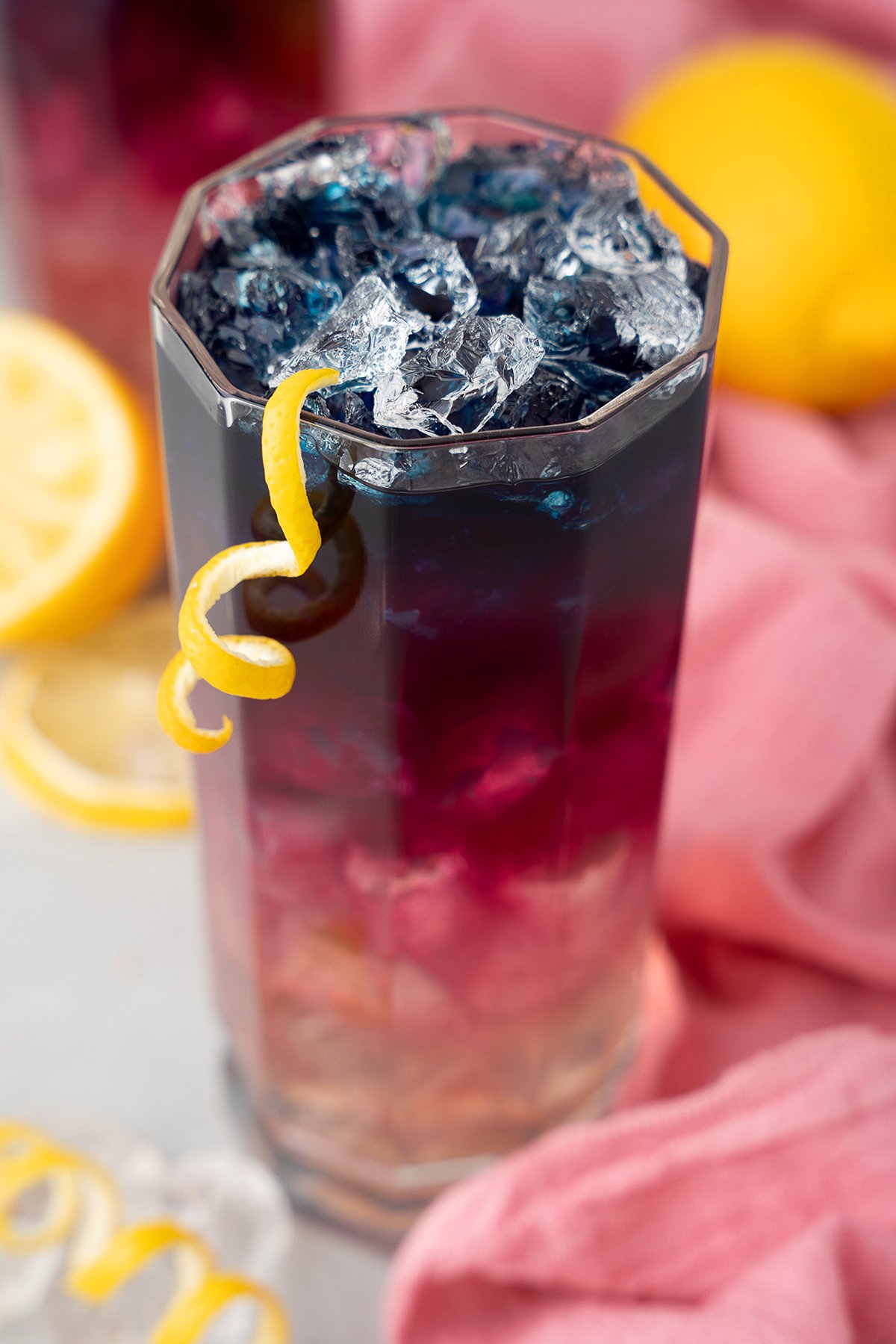 A colorful iced drink in a tall glass, garnished with a lemon twist and surrounded by lemon slices, features vibrant butterfly pea for a magical color changing lemonade experience.