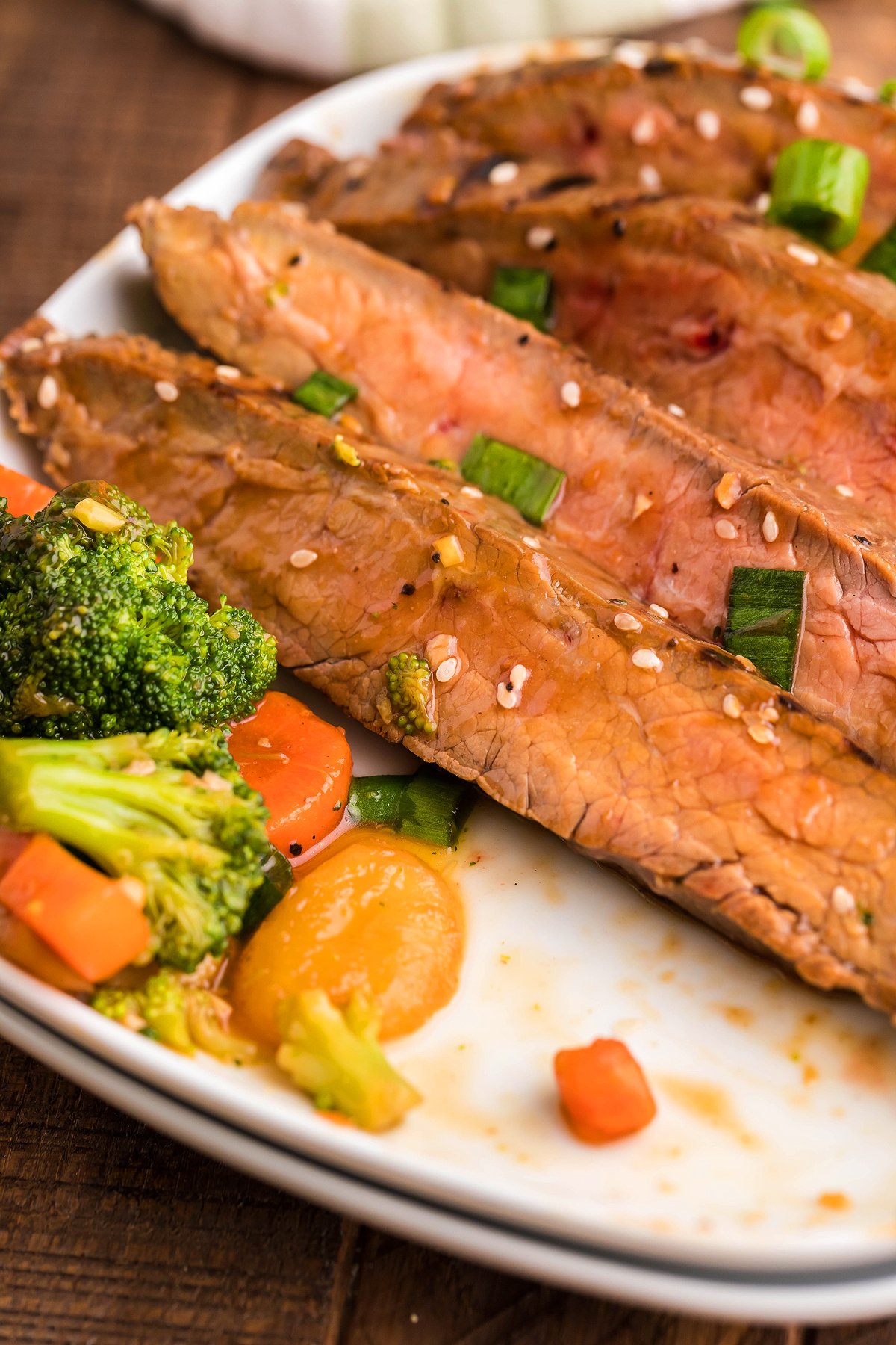 Sliced flank steak on a cast iron, topped with sesame seeds and green onions, served alongside broccoli, carrots, and squash on a white plate.
