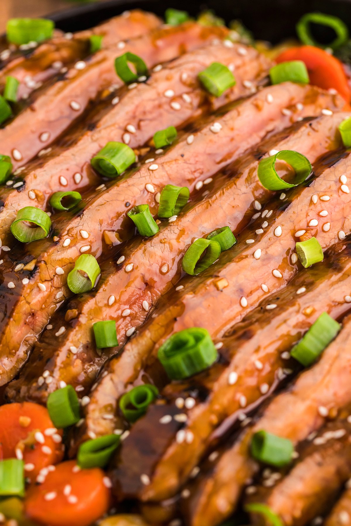 Sliced glazed beef from a flank steak recipe, topped with sesame seeds and chopped green onions, garnished with sliced carrots—perfectly seared on a cast iron grill pan for extra flavor.