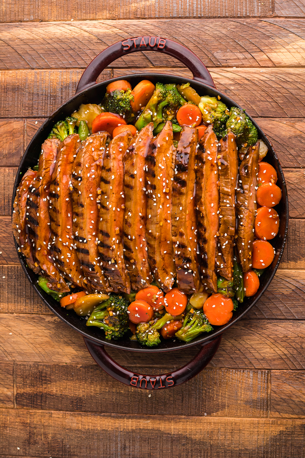 Grilled teriyaki flank steak on a cast iron, sliced over sautéed broccoli and carrots, garnished with sesame seeds in a round pan—a perfect quick steak dinner.