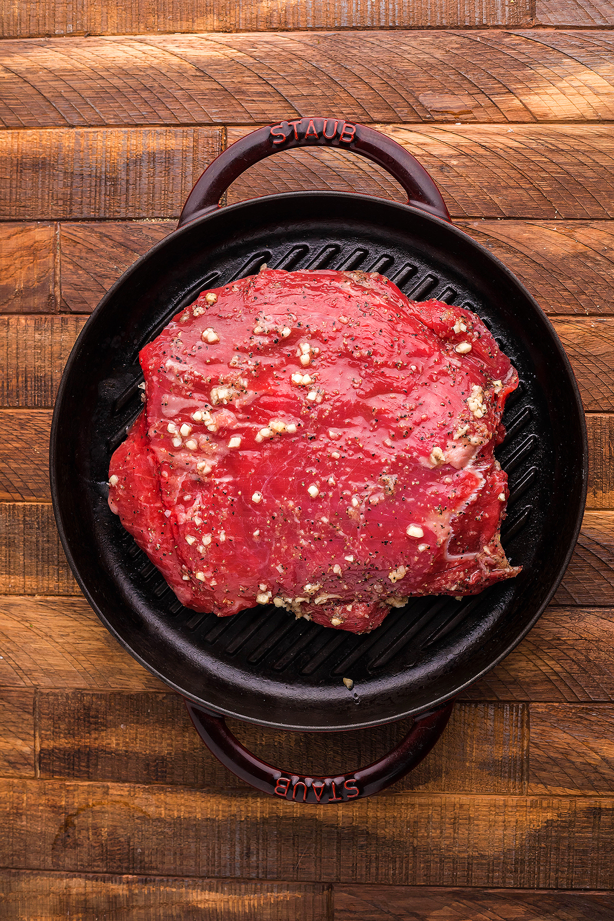 Raw marinated flank steak on a cast iron grill pan with spices sits on a wooden surface, ready to be cooked for a quick steak dinner.