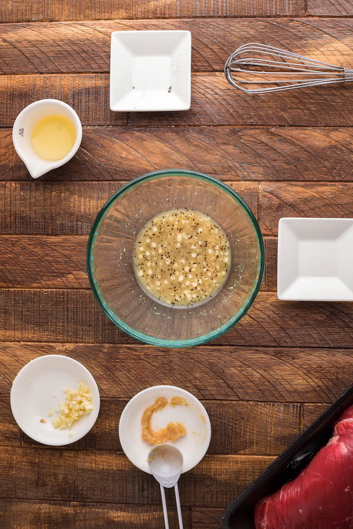 Top view of a glass bowl with seasoned mixture, perfect for a quick steak dinner, surrounded by small plates of ingredients and a whisk on a wooden table.