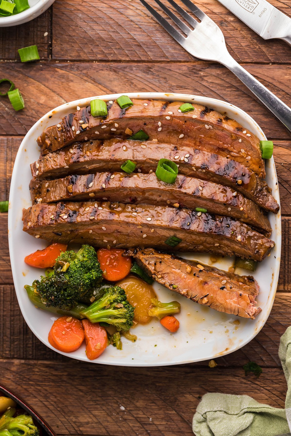 Sliced Teriyaki Flank Steak and Veggies topped with sesame seeds and green onions, served with broccoli and carrots on a white plate—a quick steak dinner perfect for any night.