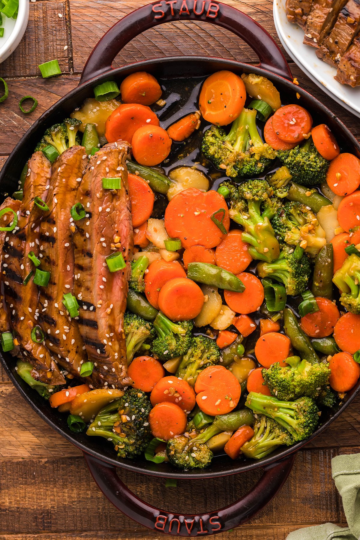 A skillet with Teriyaki Flank Steak and Veggies—sliced grilled steak, broccoli, carrots, and sesame seeds—on a wooden table.