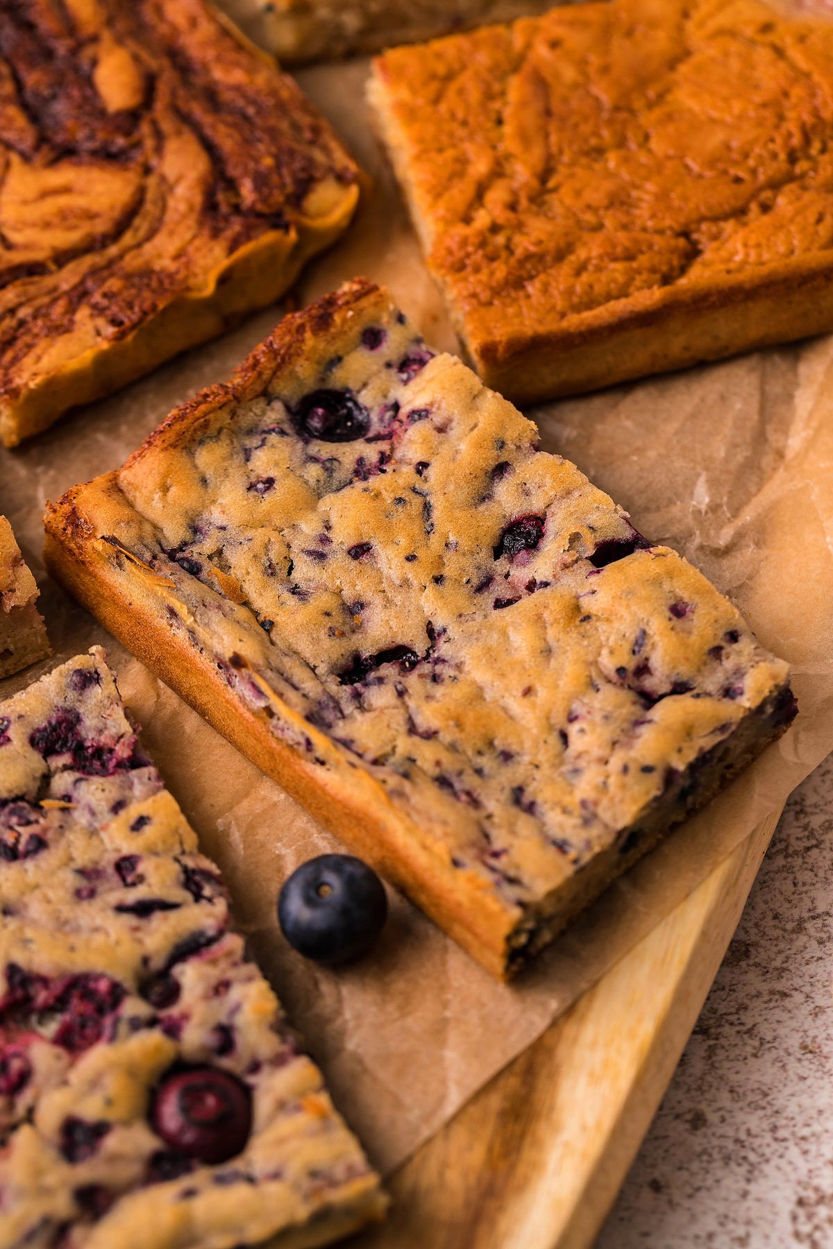 A close-up of blueberry blondies on parchment paper with a fresh blueberry beside them, reminiscent of Sheet Pan Buttermilk Pancakes—a delightful pancake hack for effortless baking.