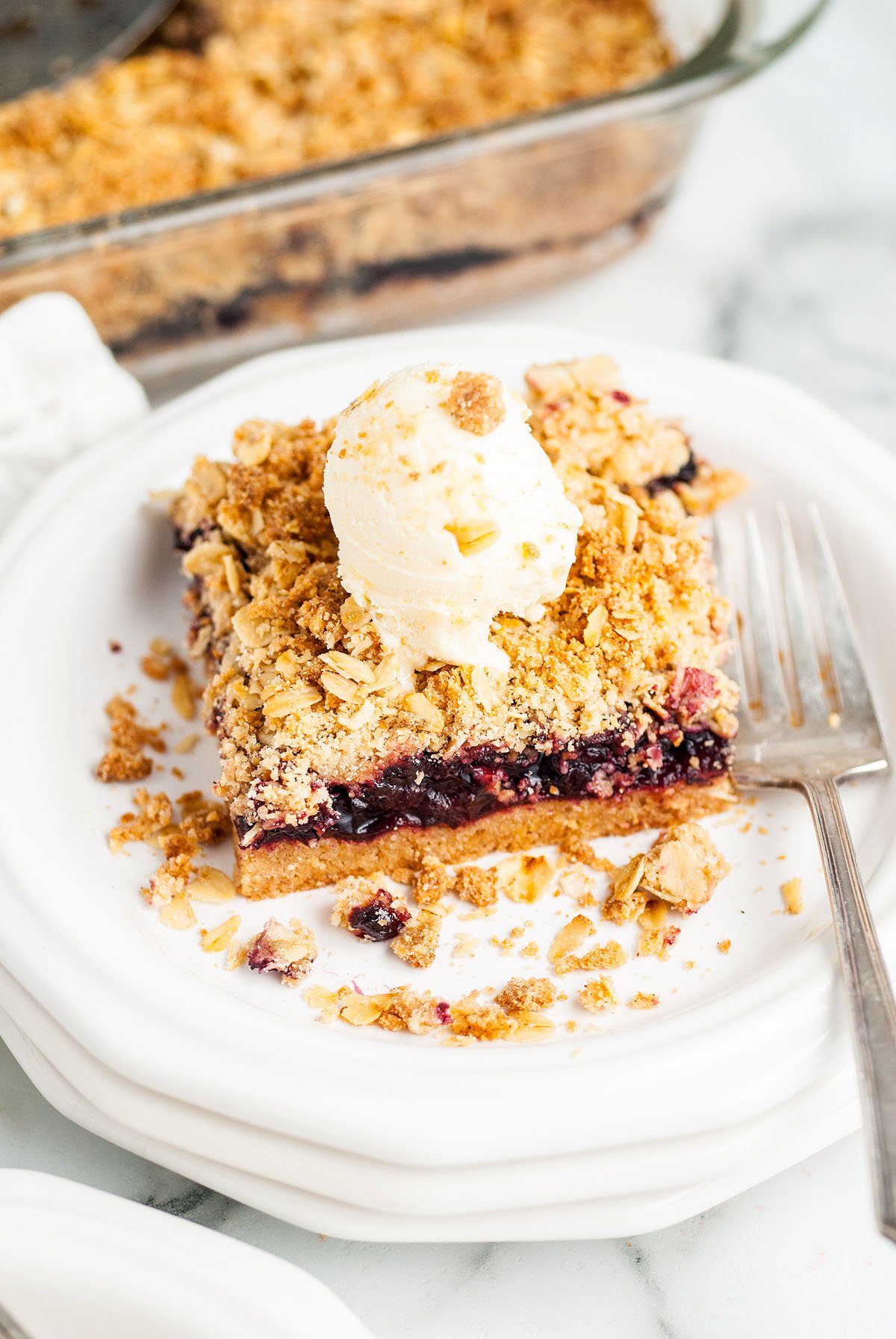 A slice of berry crumble, inspired by a Starbucks copycat recipe, topped with a scoop of vanilla ice cream on a white plate with a fork—deliciously reminiscent of oatmeal bars with cherry filling made from frozen cherries.