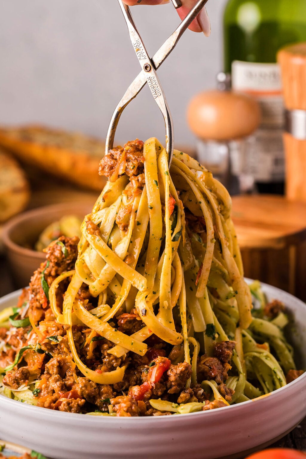 Tongs lifting pasta with meat sauce from a bowl, showcasing Homemade Italian beef bolognese, with bread and a wine bottle in the background.