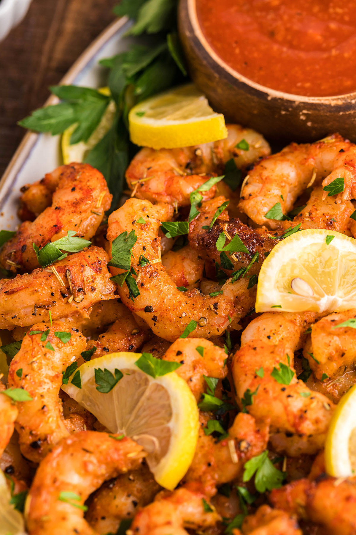 Smoked shrimp seasoned with Copycat Traeger blackened Saskatchewan rub, garnished with lemon slices and chopped parsley, served alongside a bowl of red dipping sauce.