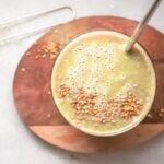 A green mango smoothie topped with seeds, served with a metal straw on a wooden board.