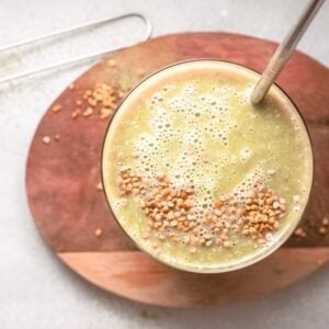 A green mango smoothie topped with seeds, served with a metal straw on a wooden board.