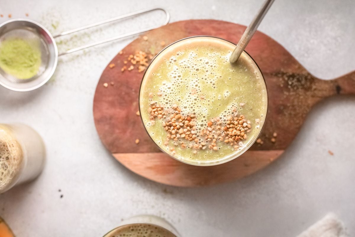 A Green smoothie in a glass with a metal straw, topped with seeds, sits on a wooden board. This Mango Matcha Smoothie blends vibrant flavors for a refreshing boost.