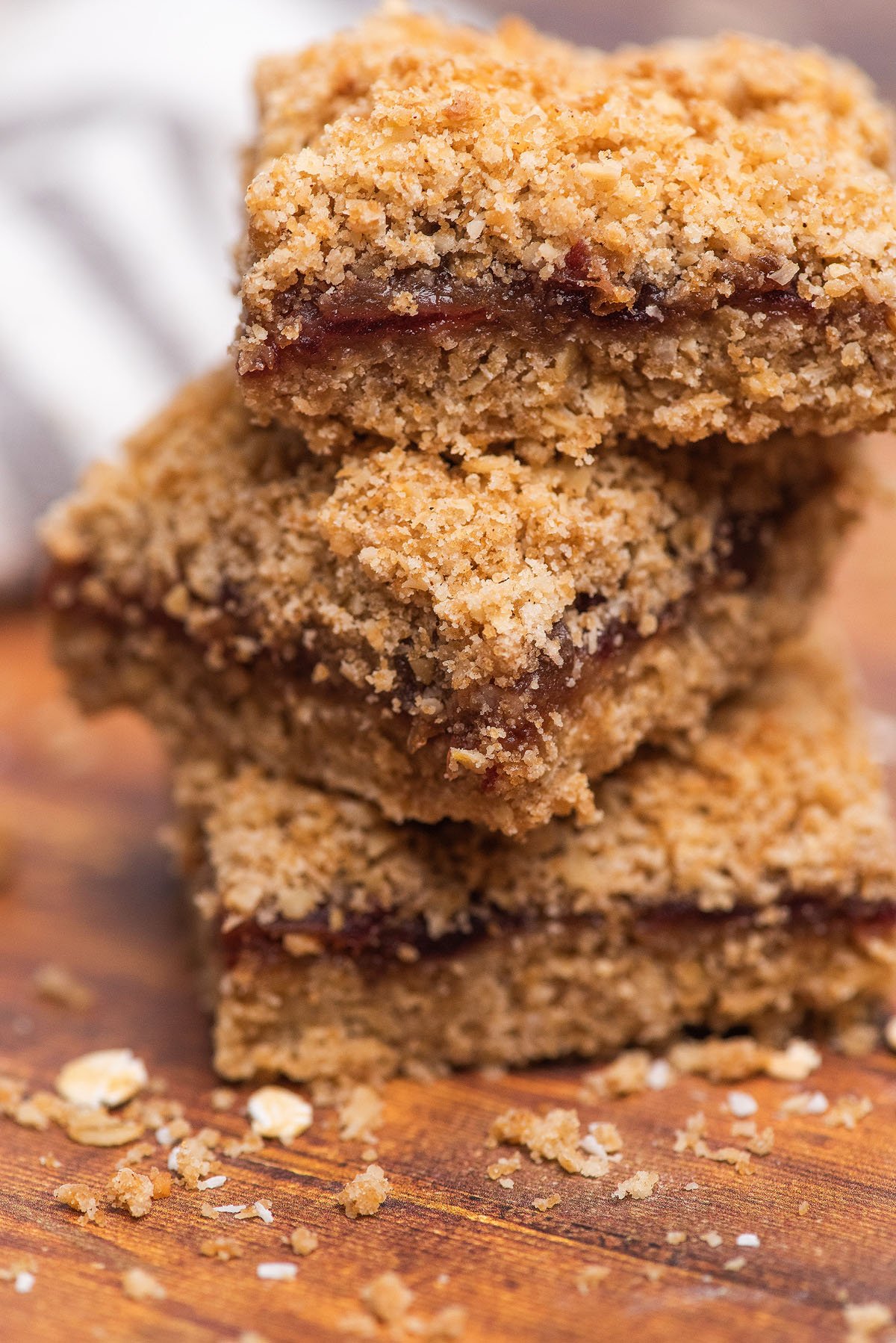 Three stacked Date Bars with a fruit filling and an Oatmeal Crumble Topping, sitting on a wooden surface with crumbs around them.