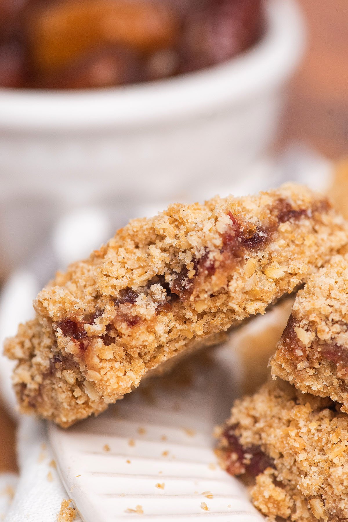 Close-up of crumbly oat bars with a sweet date crumble filling, stacked on a white plate, with a bowl in the background—perfect for anyone craving an easy recipe with dates and classic Oatmeal Crumble Topping.
