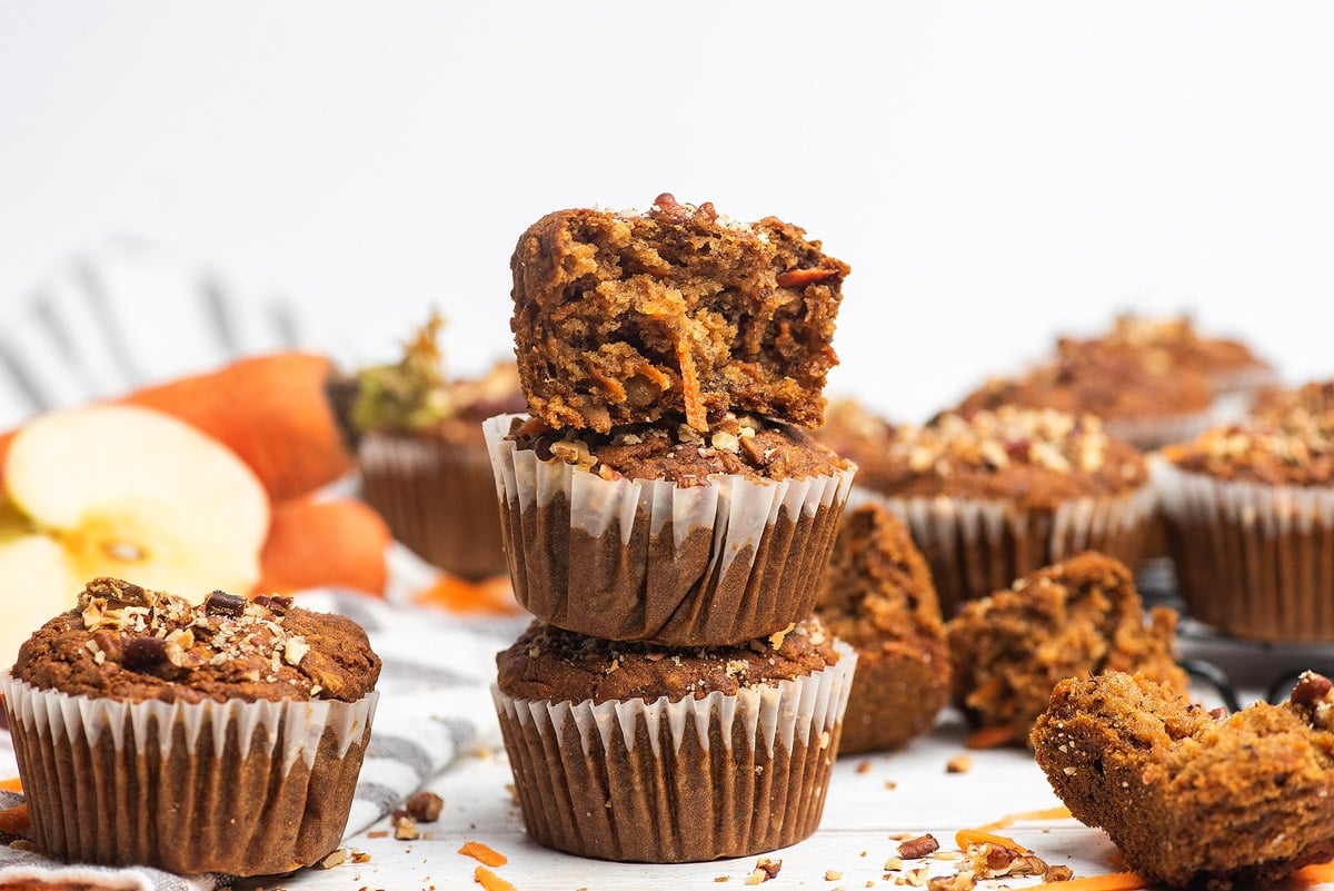 Two stacked Morning Glory Muffins, with a bite taken from the top gluten free muffin, surrounded by more muffins and apple slices.