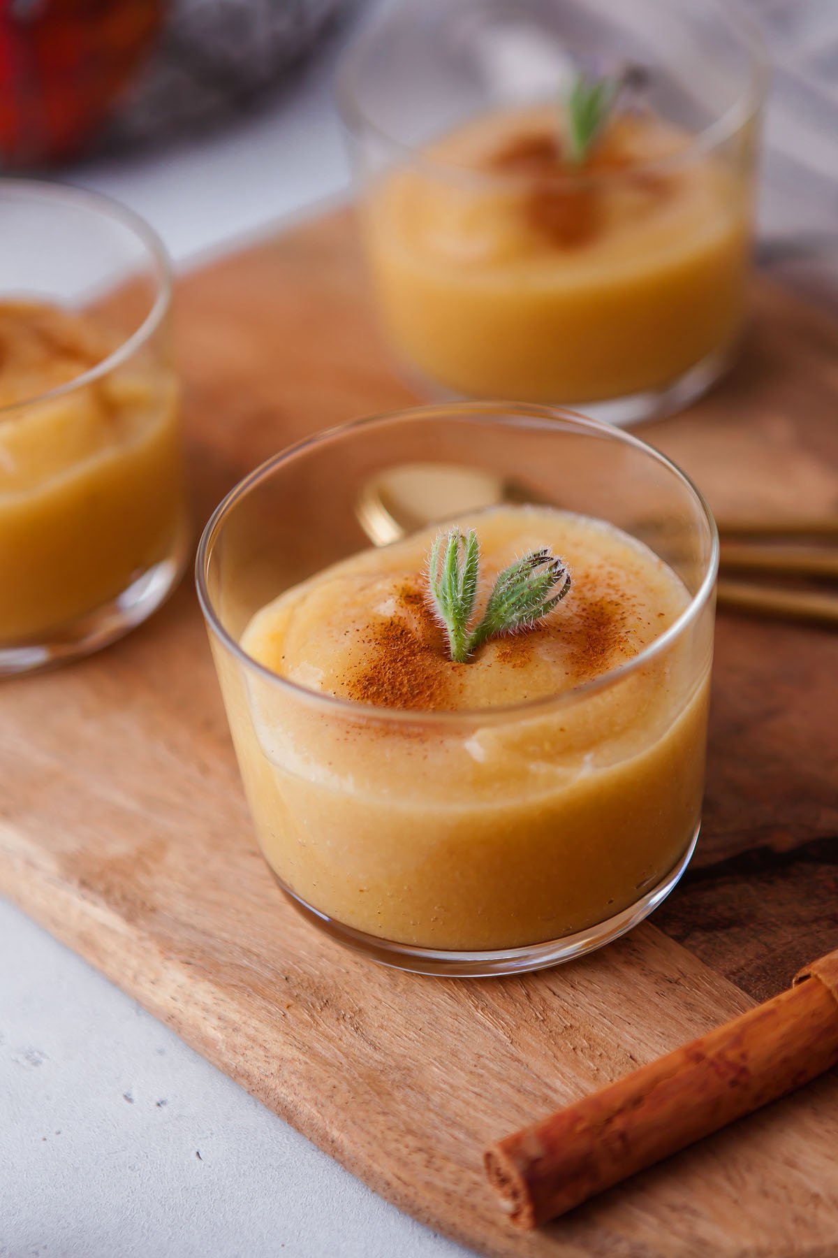A glass of creamy pudding topped with cinnamon and a green herb, placed on a wooden board near a cinnamon stick, pairs perfectly with homemade applesauce from scratch.