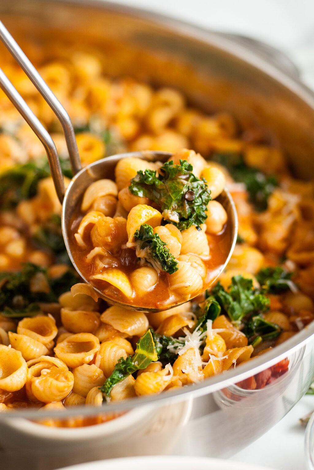 A ladle scooping shell pasta with tomato sauce and kale from a pan.