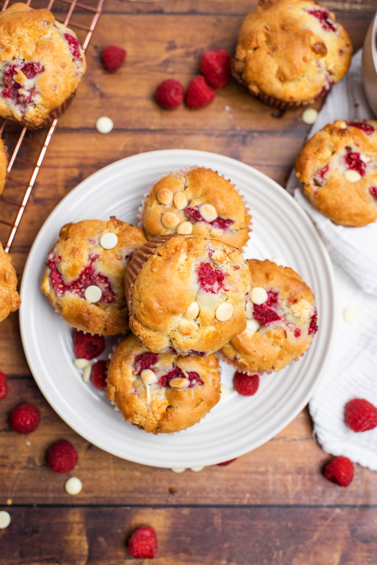 A plate of freshly baked raspberry white chocolate muffins sits on a wooden table, surrounded by scattered berries and chocolate chips.