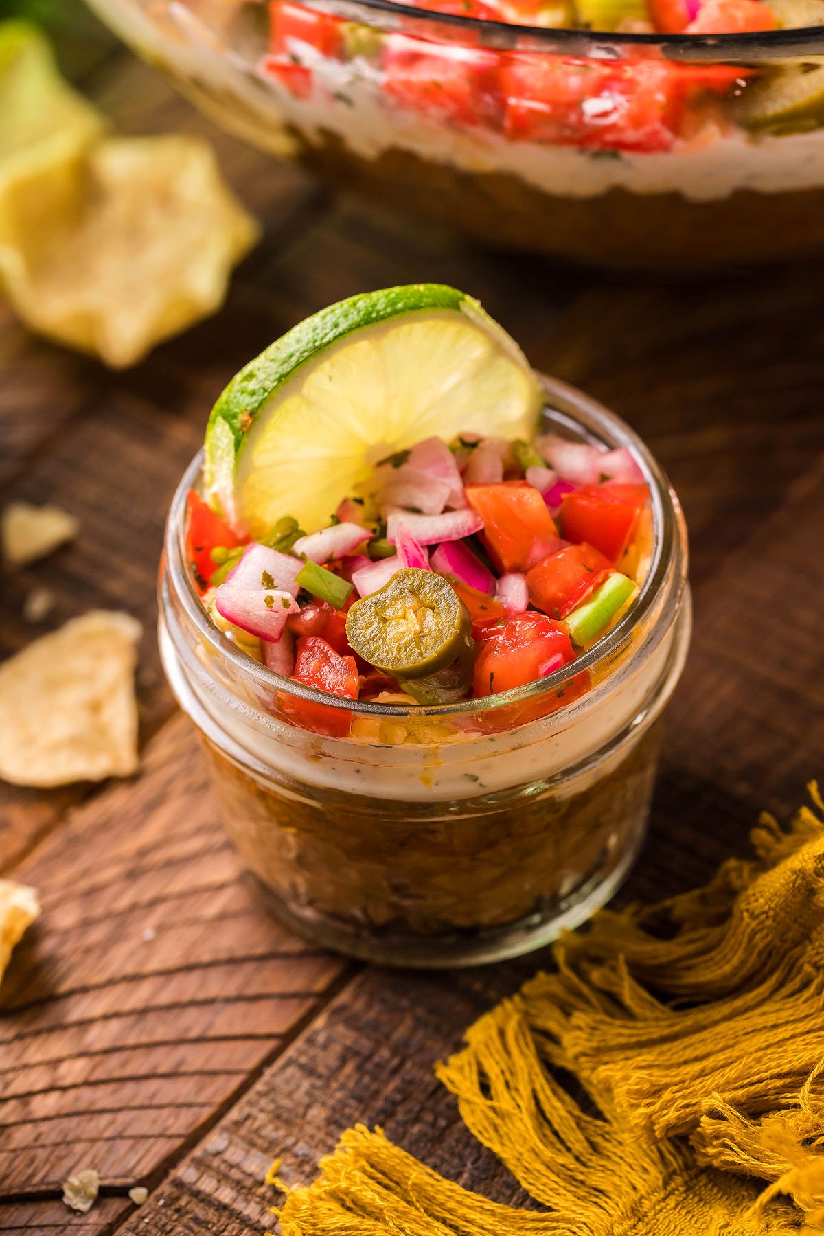 A jar of 5 Layer Taco Dip topped with diced veggies, jalapeño, and a lime slice sits on a wooden table—an easy appetizer perfect for any occasion.