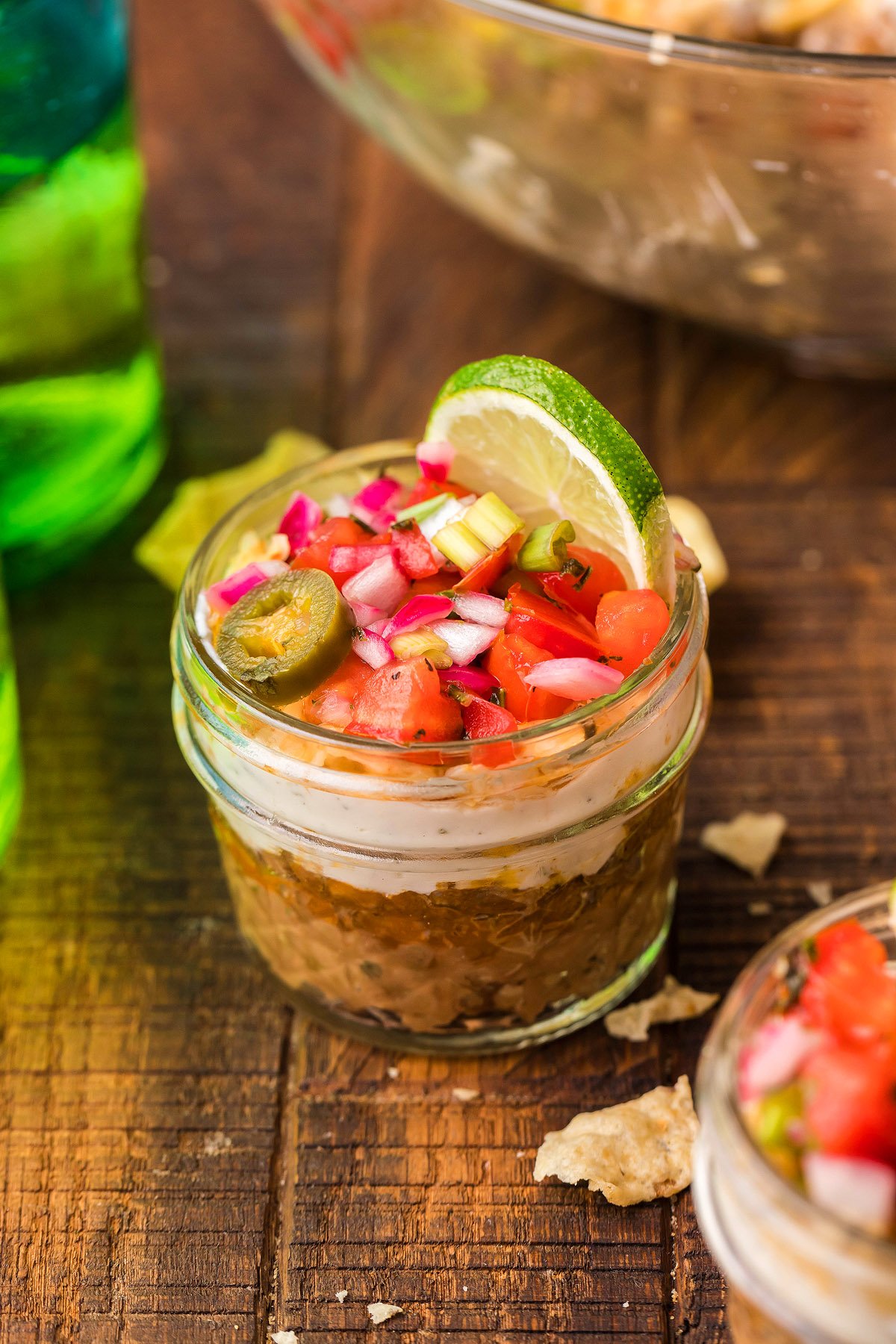 Small glass jar filled with 5 Layer Taco Dip, topped with chopped veggies, jalapeño, and a lime slice—an easy appetizer perfect for any Mexican layered dip lover.