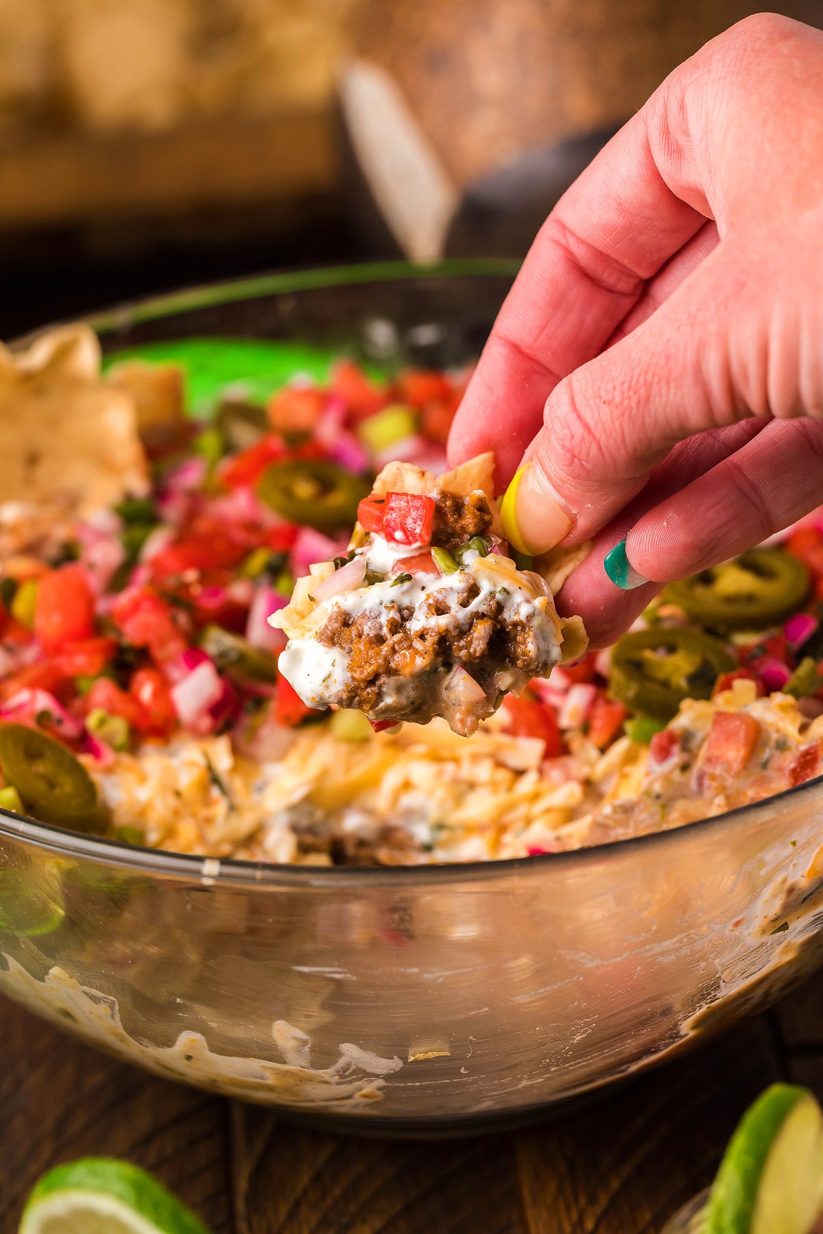 A hand holds a chip with layered taco dip above a bowl filled with colorful 5 Layer Taco Dip, a delicious Mexican layered dip perfect for parties.