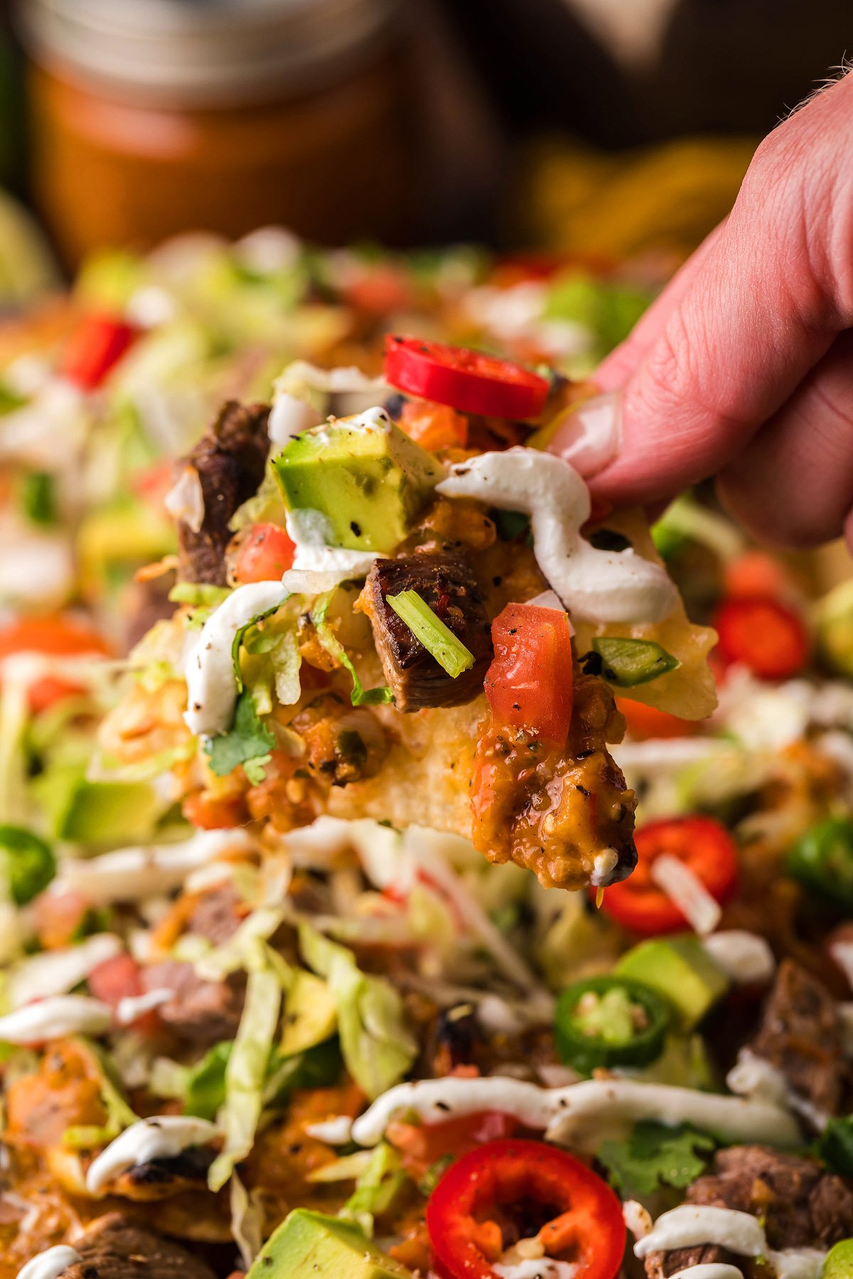 A hand holding a loaded nacho chip with beef, avocado, tomatoes, cheese, sour cream, and classic nacho toppings—perfect for Mexican steak nachos.