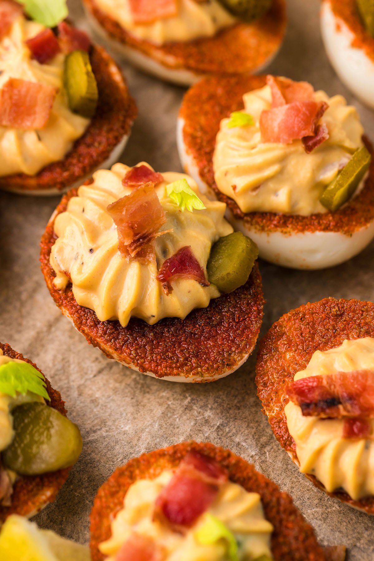 Unique gourmet deviled eggs topped with pickles, bacon bits, and herbs are artfully arranged on a parchment-lined surface.