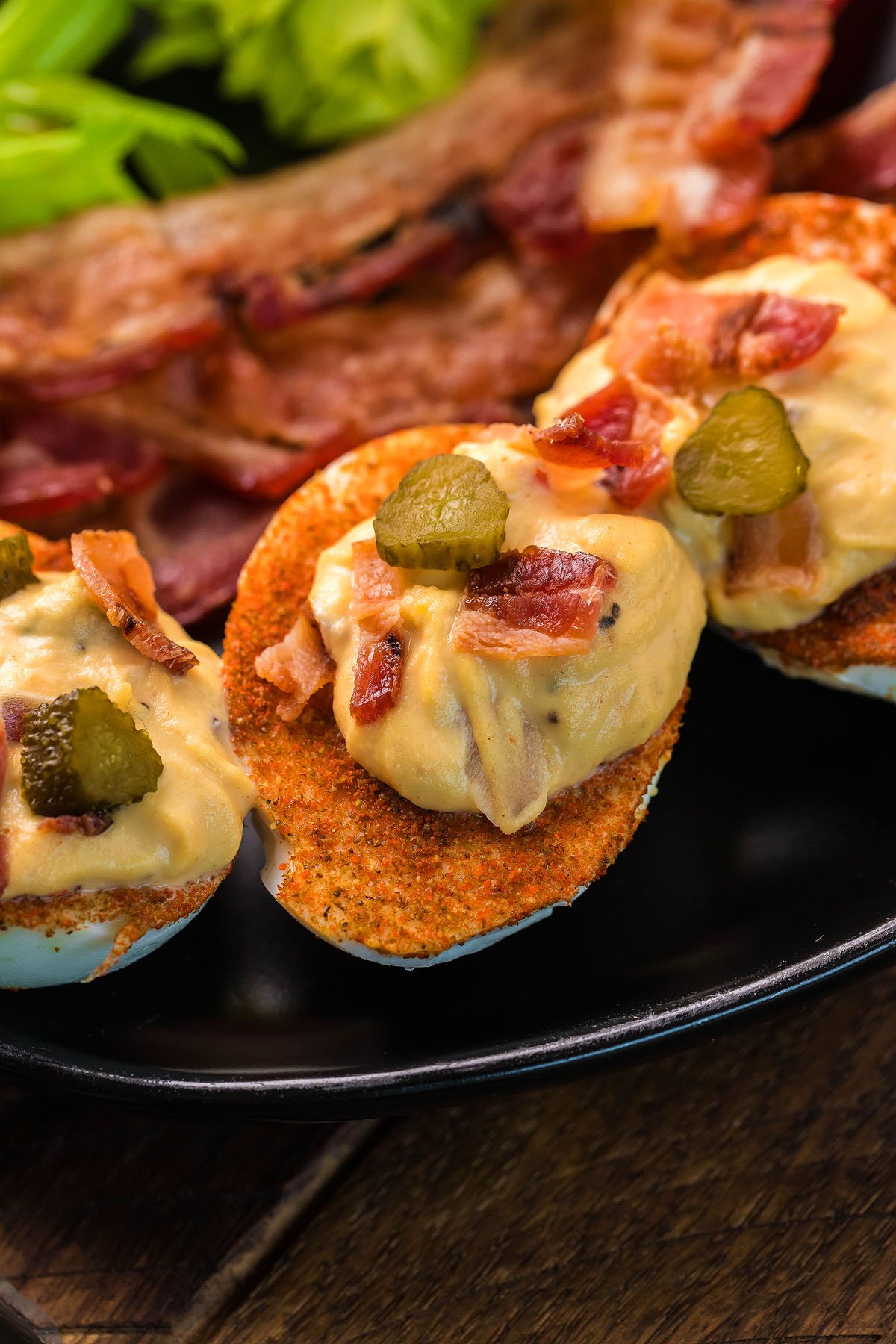 Unique gourmet deviled eggs topped with crispy bacon pieces and pickle slices, these loaded deviled eggs are served on a sleek black plate.