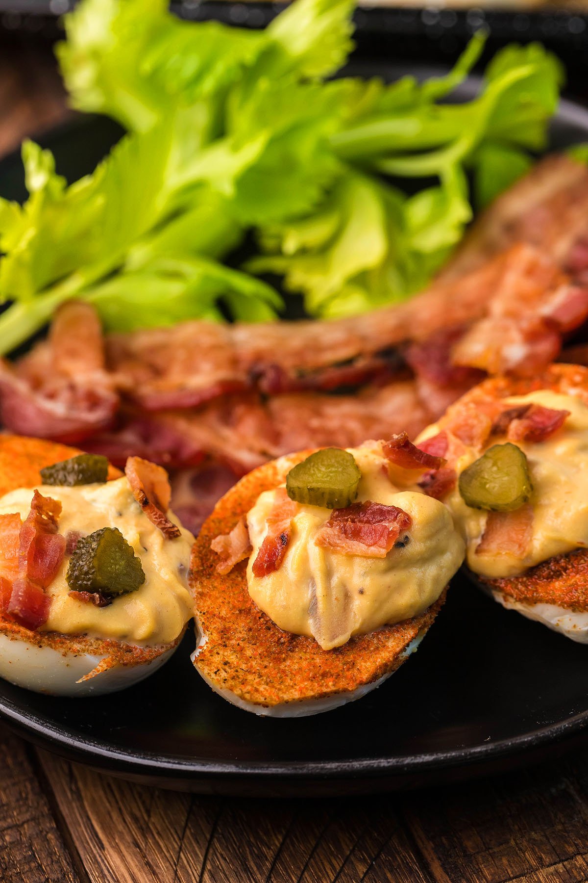 Loaded deviled eggs topped with bacon and pickles, served with celery and extra bacon strips on a black plate—a unique gourmet deviled egg experience.