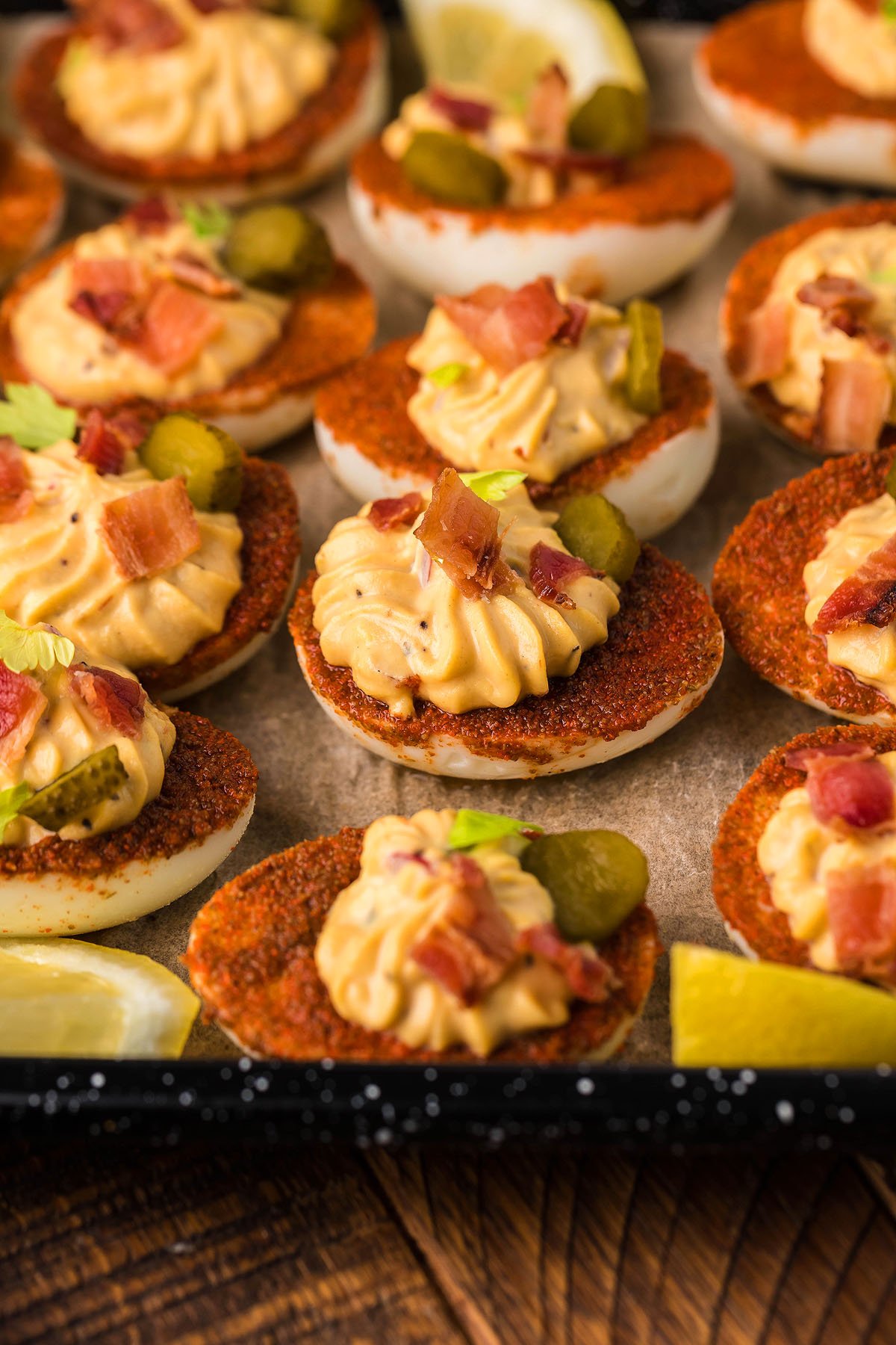 These loaded deviled eggs are topped with paprika, bacon bits, pickles, and herbs on a tray with lemon wedges—a creative deviled egg idea perfect for any gathering.