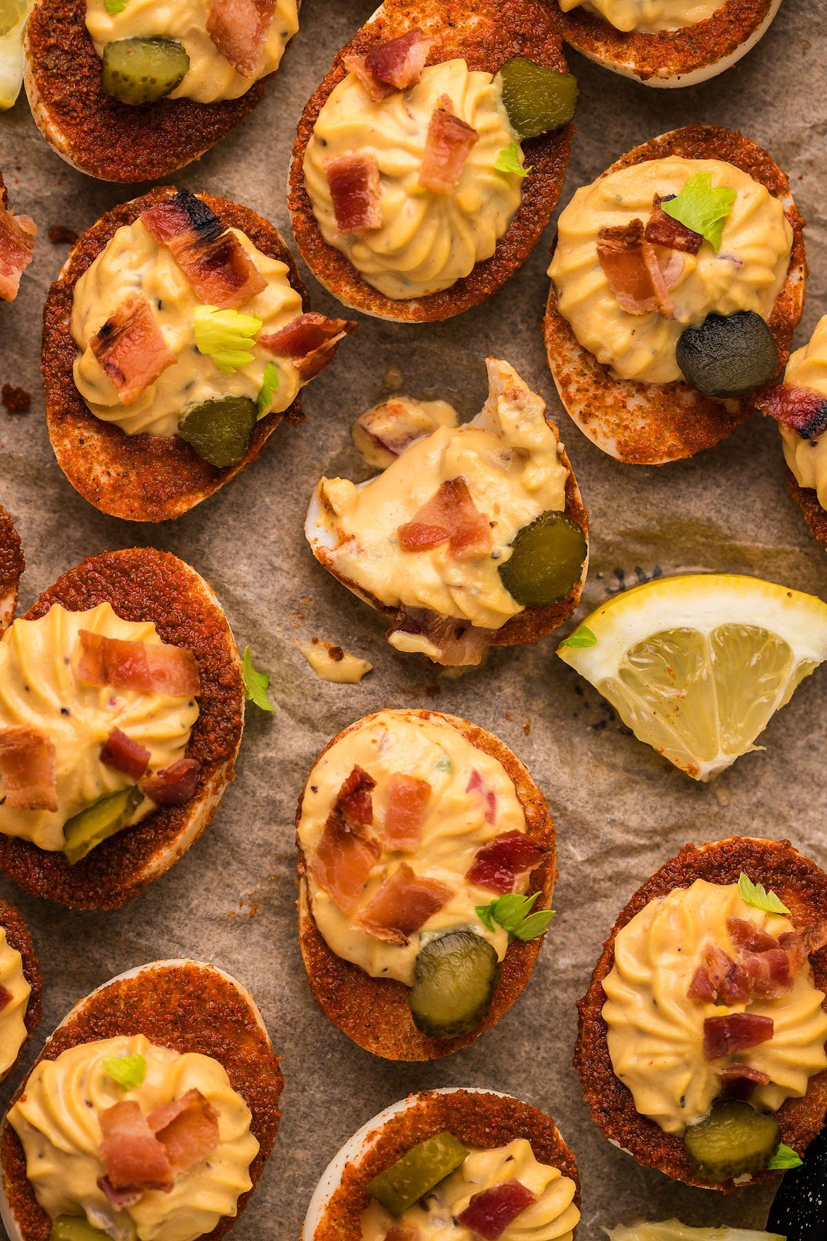 A tray of loaded deviled eggs topped with bacon, pickles, and herbs, served with a lemon wedge on the side.
