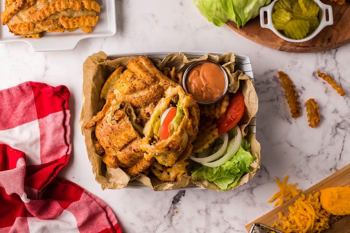 A fried chicken sandwich with lettuce, tomato, pickles, and sauce, served with waffle fries and a tasty French Fry Topping on parchment.