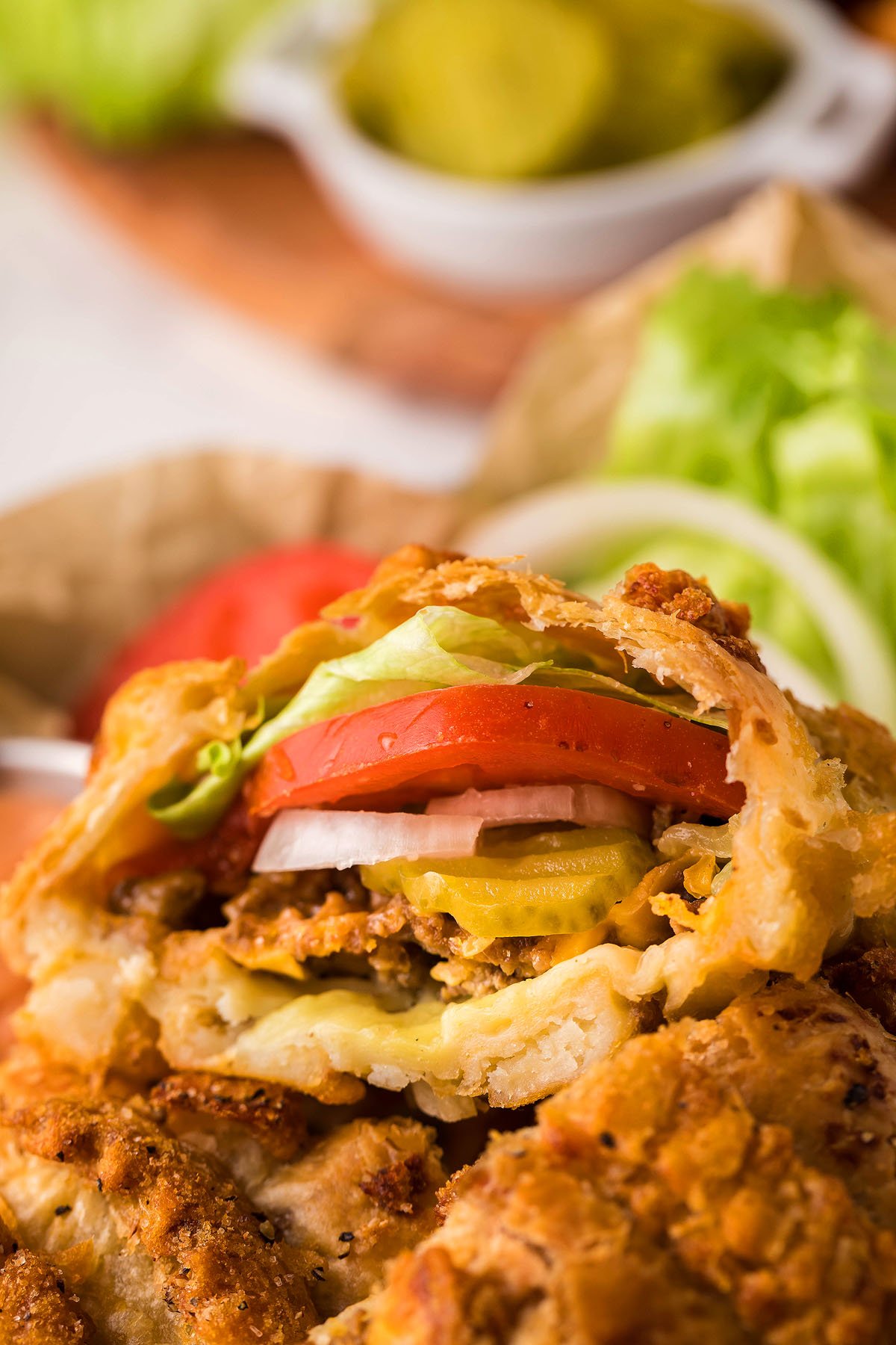 Close-up of a crispy fried sandwich inspired by Cheeseburger hand pies, filled with lettuce, tomato, onion, and pickles, with fresh toppings in the background.