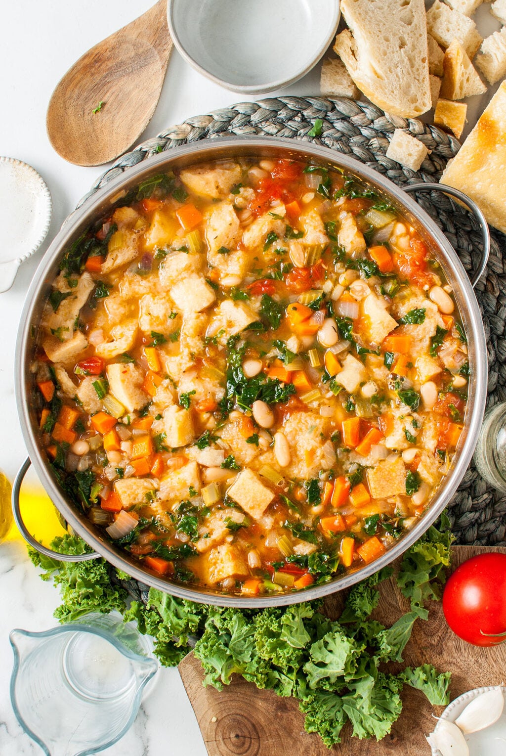 A pot of Ribollita - Italian Bean Soup with beans, bread pieces, carrots, and greens sits on a woven mat, surrounded by bread and fresh greens.