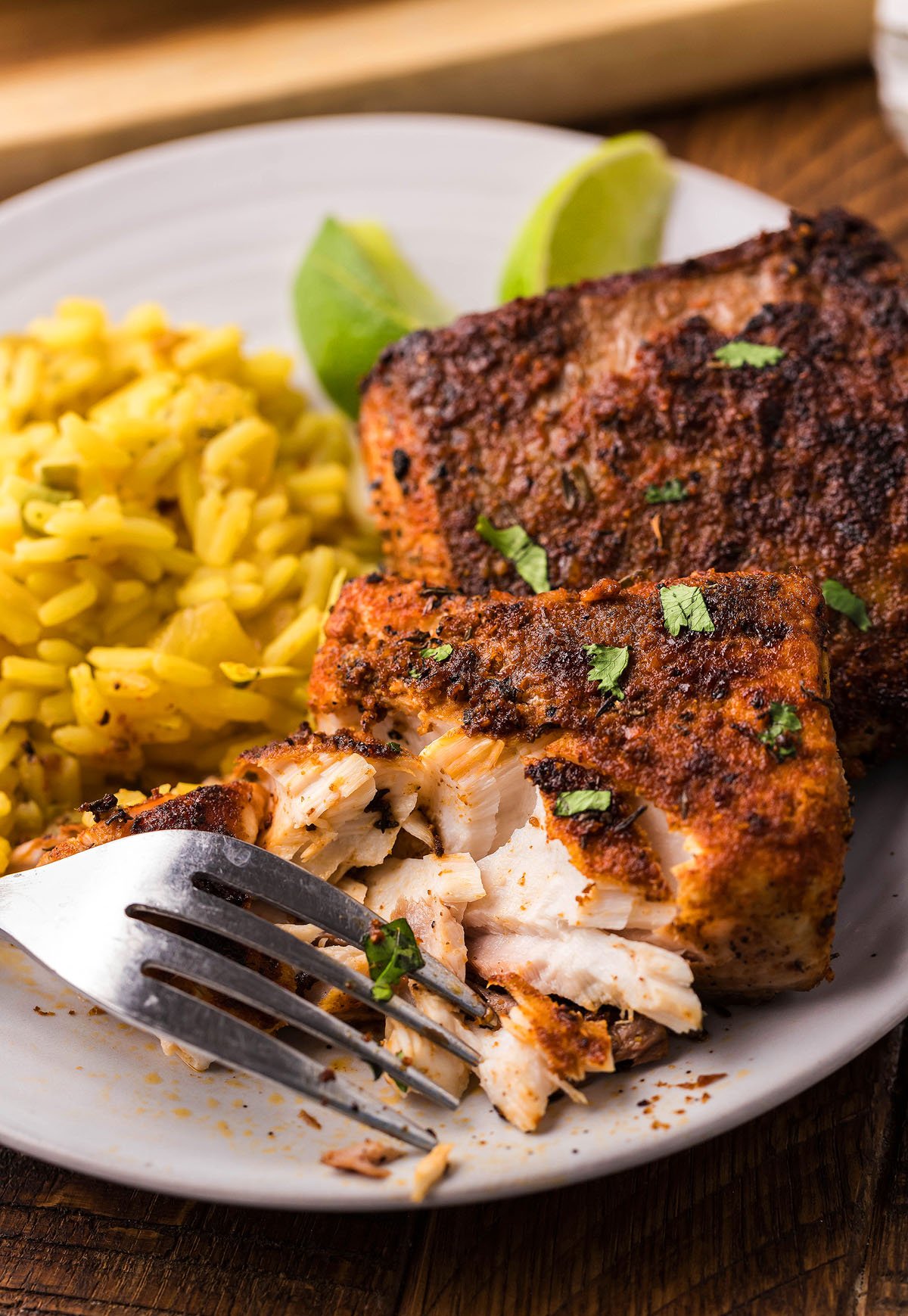 A fork flakes Blackened Mahi Mahi, seasoned with homemade seasoning, alongside yellow rice and lime wedges on a plate.