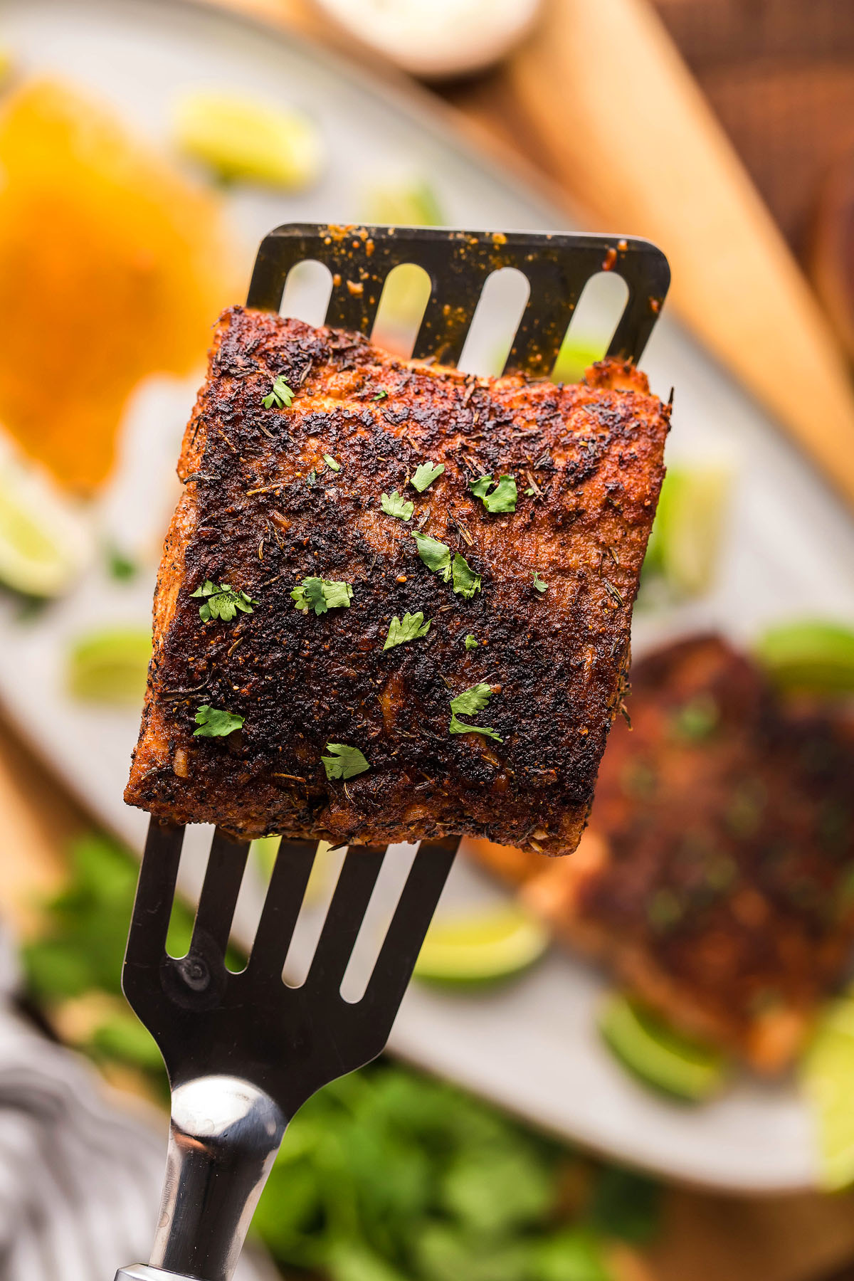 A metal spatula holds a Cajun seasoned fish fillet, blackened to perfection in a cast iron skillet and garnished with fresh herbs.