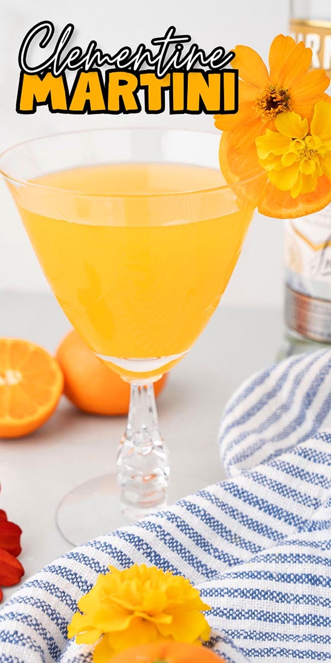 A clementine martini in a glass, garnished with an orange flower, with clementines and a striped cloth nearby.